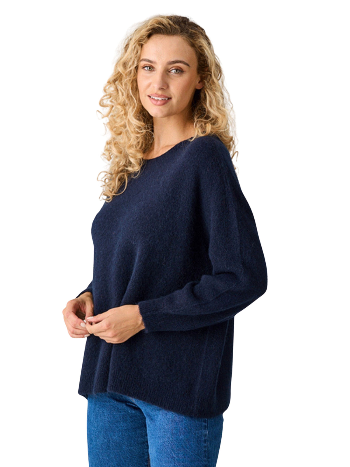 A woman with curly blonde hair wearing a dark navy blue knit sweater and blue jeans, standing against a plain white background.