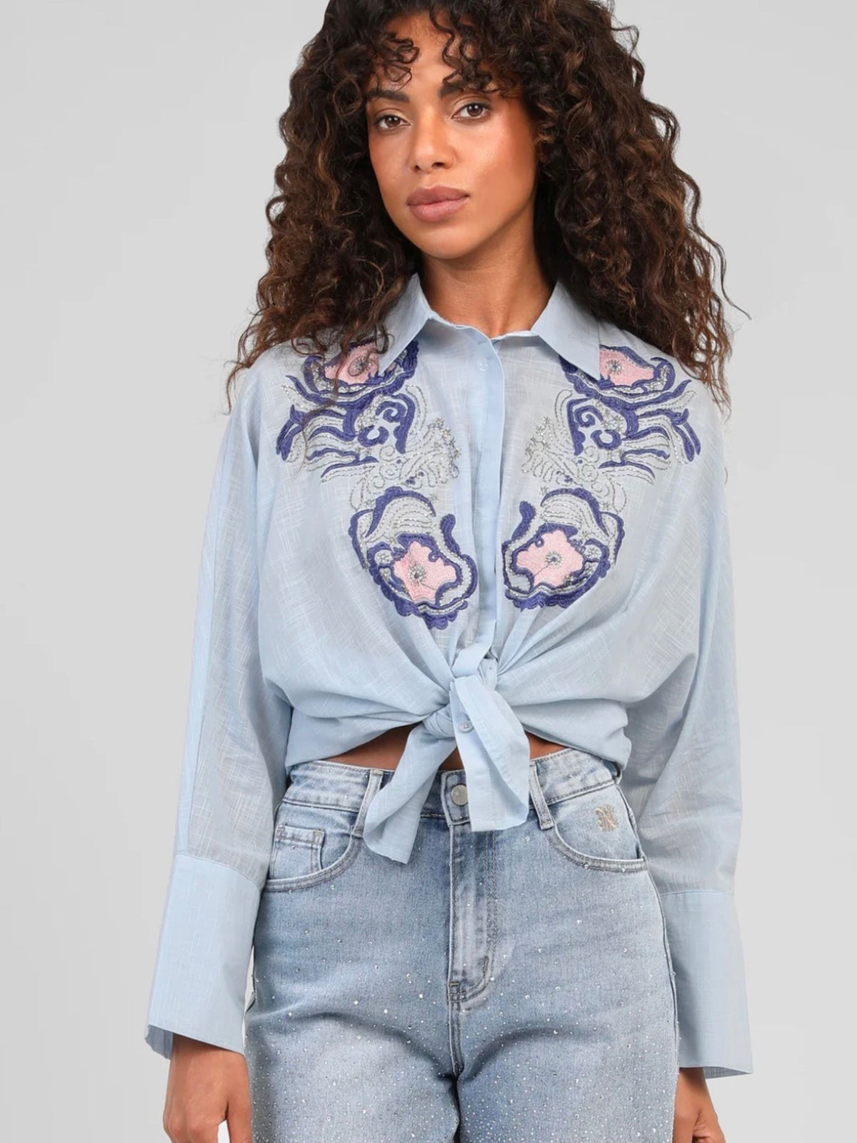 A woman wearing a light blue long-sleeve button-up shirt with purple and pink floral embroidery across the chest, tied at the waist, paired with light-wash jeans against a plain light gray background.